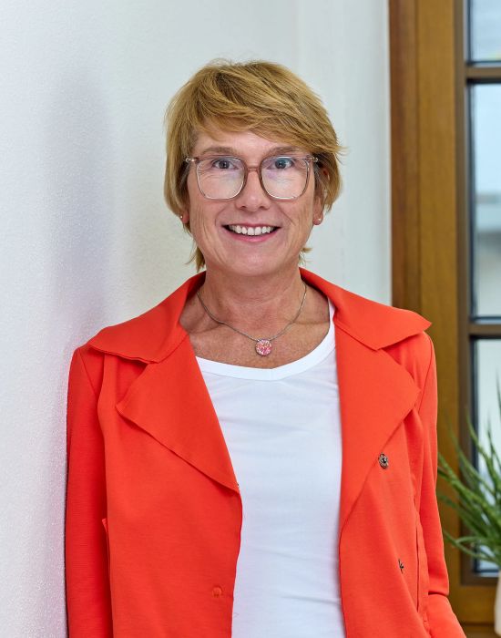 Frau mit kurzer blonder Frisur mit Brille rotem Blazer und weißem T Shirt laechelt freundlich