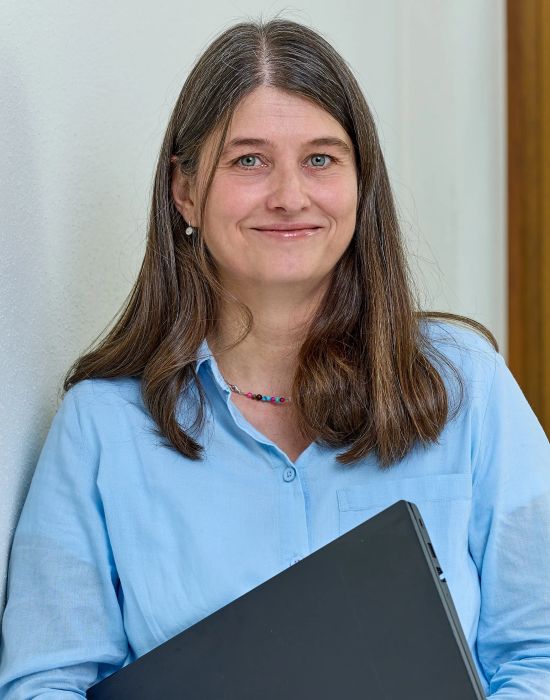 Frau mit blauer Bluse und Laptop vor Fenster mit Pflanzen im Hintergrund