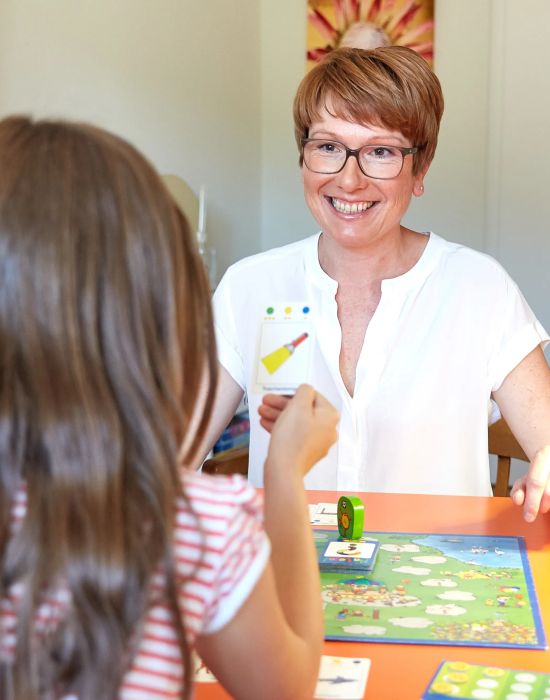 Logopaedin und Kind spielen gemeinsames Spiel am orangenen Tisch in Logopaediepraxis