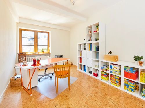 Helles Buerozimmer mit Arbeittisch Fenster und Regal mit Spielen und Ordnern